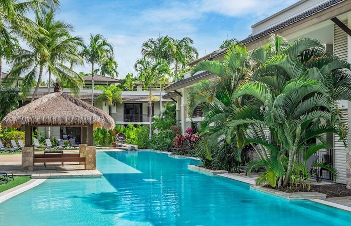 A tropical oasis is presented with a lagoon pool surrounded by lush palm trees and vibrant greenery. A thatched gazebo provides shade nearby, while lounge chairs are positioned along the poolside, inviting relaxation under the clear blue sky.