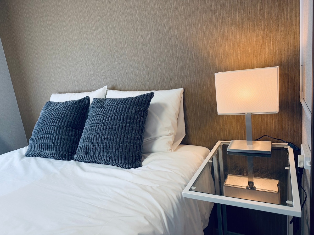 A cozy sleeping area featuring a spacious bed adorned with two textured gray pillows. A neatly arranged bedside table with a glass top supports a soft-lit lamp, contributing to a serene atmosphere. The wall is covered in a subtle, textured pattern.