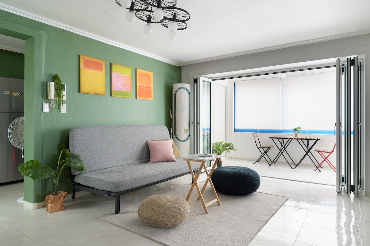 A cozy living area is highlighted by a gray sofa and a wooden side table. Green walls complement minimalist décor, with three framed artworks enhancing the space. Sliding glass doors open to a bright dining area, furnished with a small table and chairs.