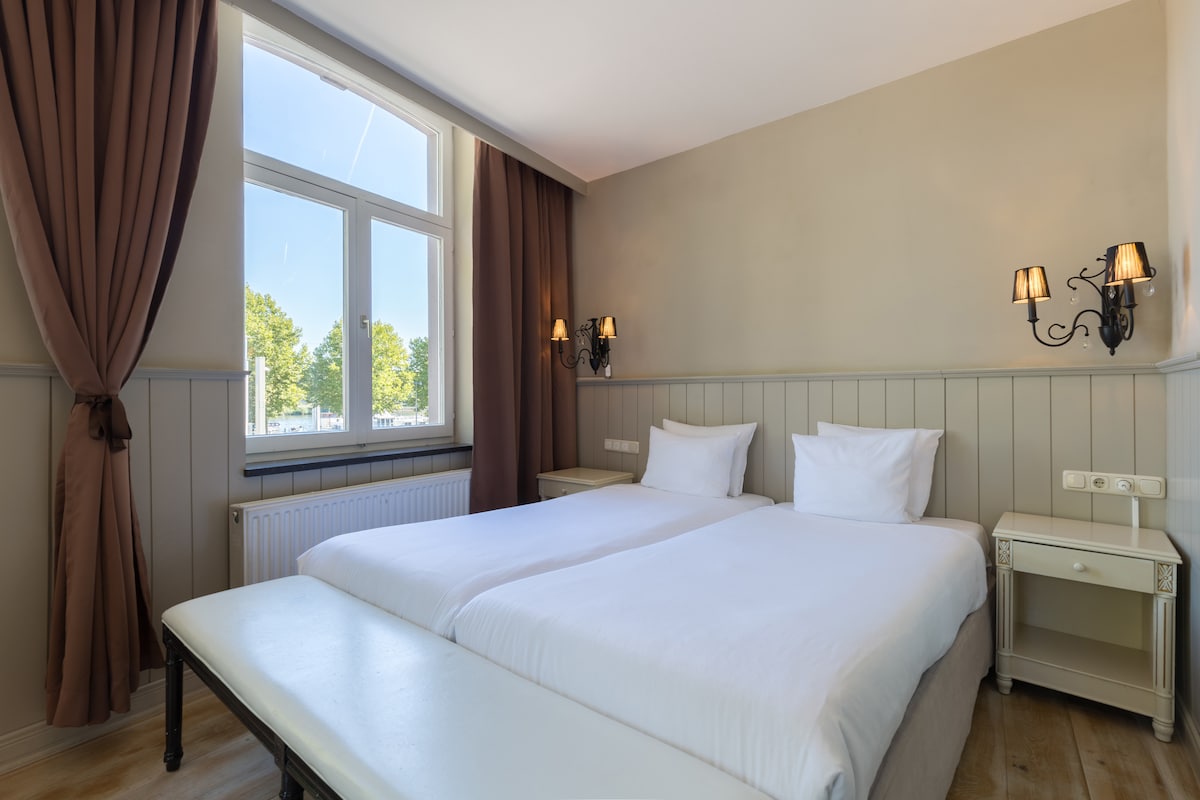 A well-lit room features two spacious beds positioned side by side, each topped with white linens and soft pillows. Natural light enters through a large window, which is framed by elegant curtains. Wall sconces add a touch of character beside the beds.