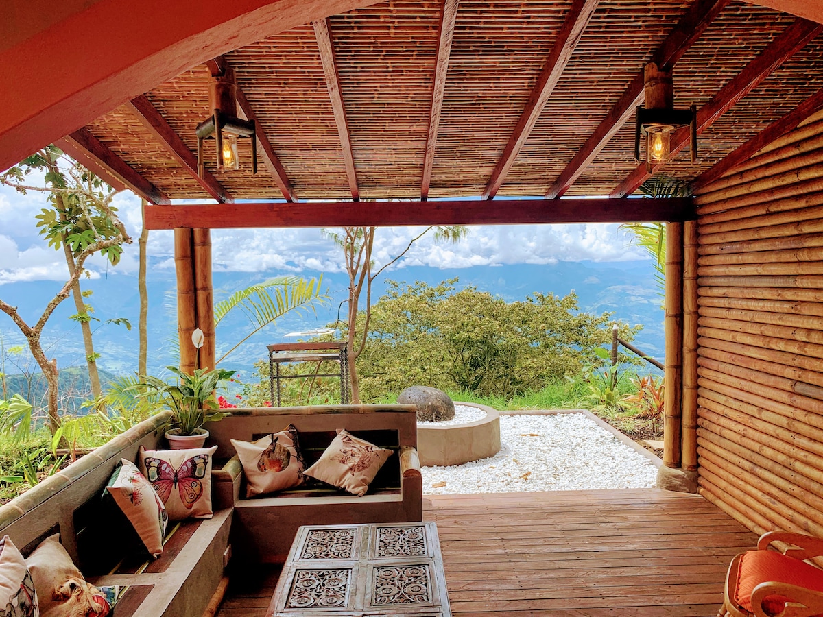 A spacious outdoor seating area features comfortable couches adorned with decorative pillows, overlooking a vast landscape. The wooden structure with a bamboo ceiling allows natural light to filter through. A small table sits center, with a stone circle and greenery in the background, enhancing the serene ambiance.