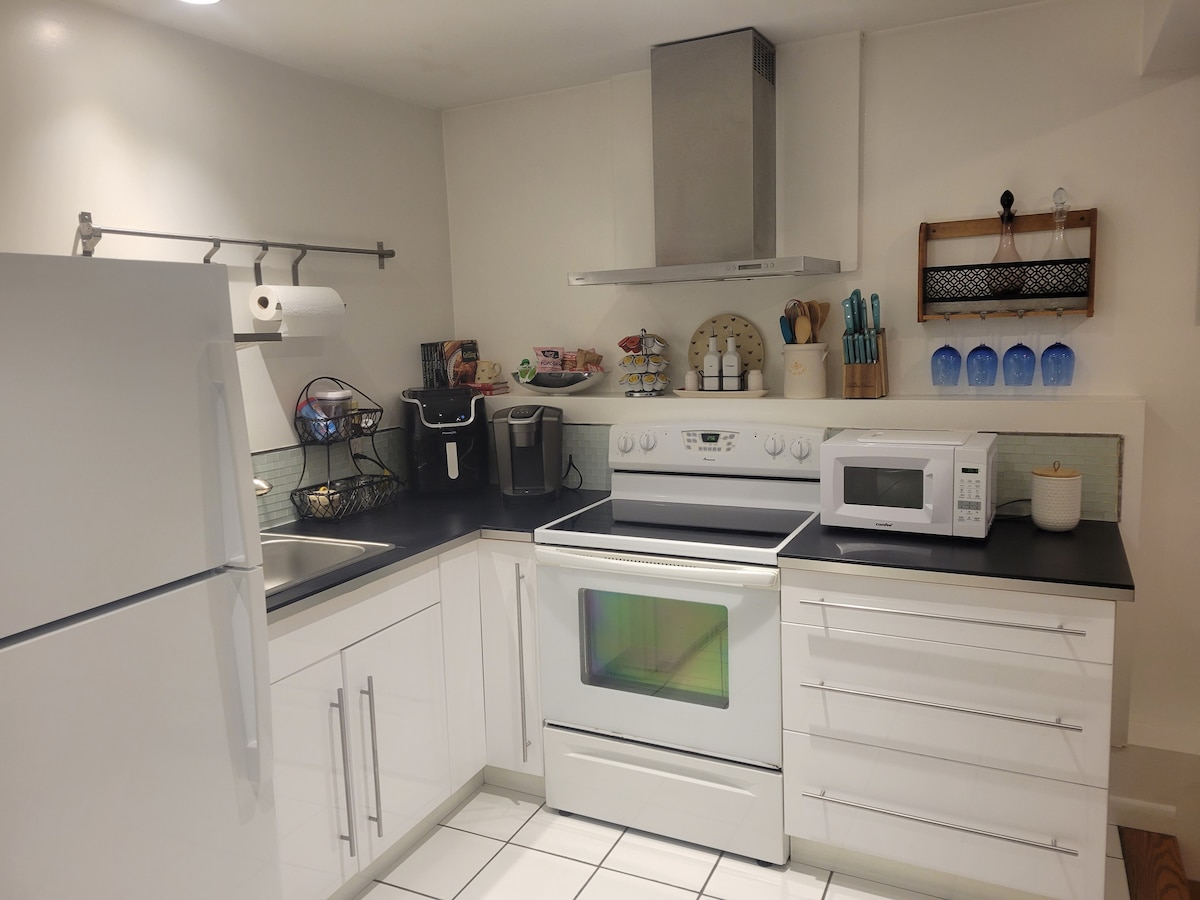 A modern kitchen is featured, equipped with a white refrigerator, oven, and microwave. A sleek sink is located near a black countertop. Cookware and utensils are neatly arranged on a wall-mounted rack, while colorful glassware is displayed on a shelf, enhancing the organized appearance.