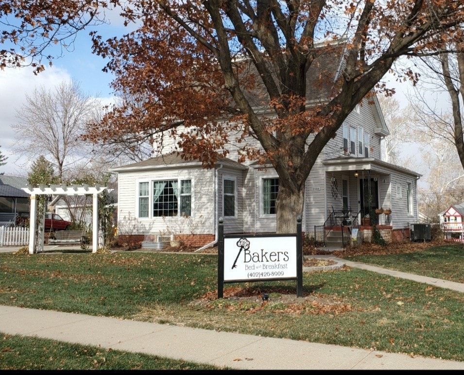 The exterior of a traditional farmhouse is featured, showcasing a corner property with a sign reading 'Bakers Bed & Breakfast.' A pergola stands beside a green lawn, and large windows are visible, allowing natural light. The surrounding trees display autumn leaves.