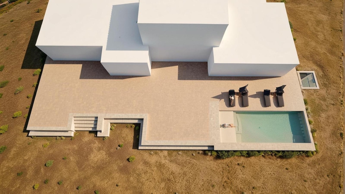 An aerial view of the villa showcases a spacious outdoor area featuring a private swimming pool. The surrounding terrain is minimally landscaped with patches of greenery. Black lounge chairs are arranged near the pool, and the villa's modern white architecture is prominently displayed.