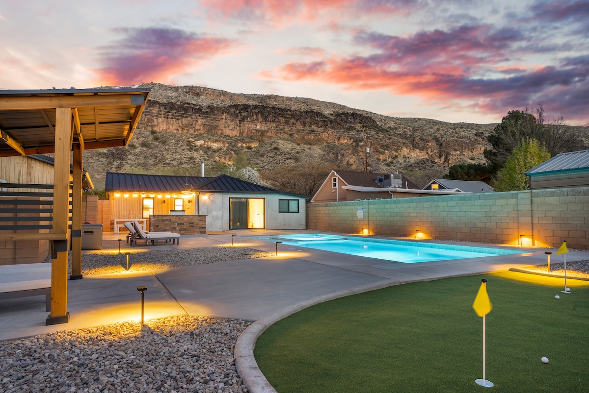An open outdoor area features a swimming pool illuminated by soft lighting, set against a backdrop of rugged mountains. A well-maintained lawn with putting greens and lounge chairs adds to the inviting atmosphere, while a modern structure is visible in the distance.