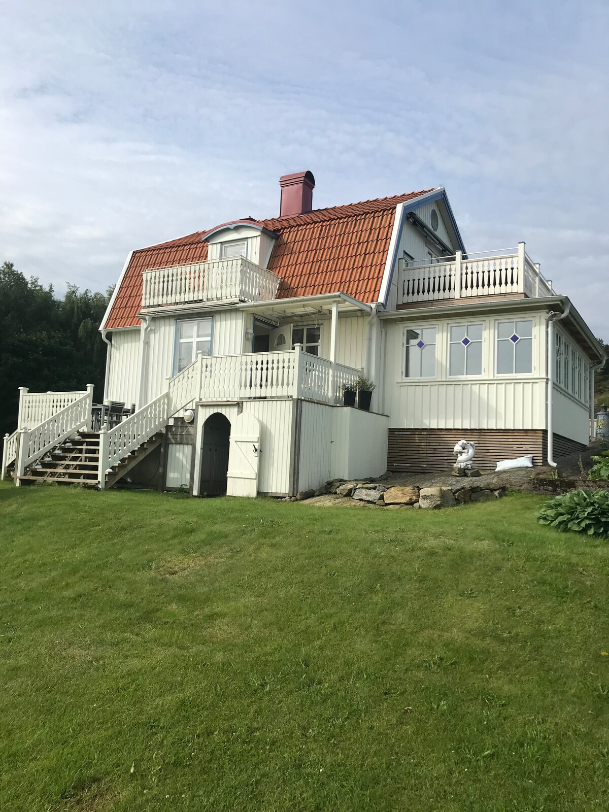 A white, three-story house features a red-tiled roof and several balconies. The structure is surrounded by a green lawn, with stairs leading to the main entrance. Large windows offer views of the outdoors, contributing to a bright and airy appearance.
