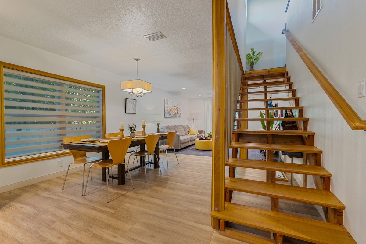 An open dining area is visible, featuring a wooden table surrounded by six light wood chairs. Natural light filters through a large window with layered shades. A staircase with wooden steps leads up, while a cozy living area is seen in the background.