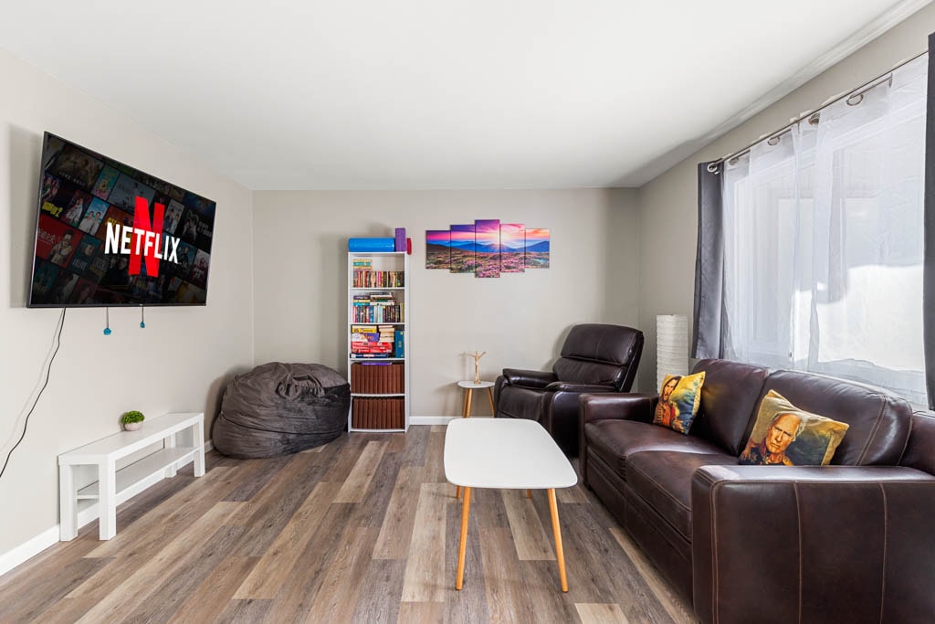 A living room space features a large flat-screen television mounted on the wall, displaying the Netflix logo. Dark leather seating is arranged around a white coffee table, with a bean bag chair nearby. Colorful wall art adds visual interest, while natural light filters in through the windows.