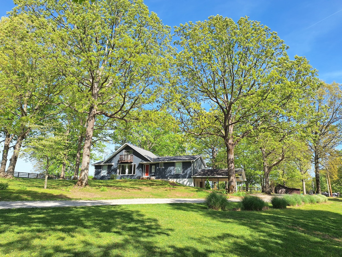 A spacious country home is set among lush green trees, with well-maintained grass surrounding the property. The blue sky provides a bright backdrop, while large windows reflect natural light. The inviting entrance features a pathway leading to the house.