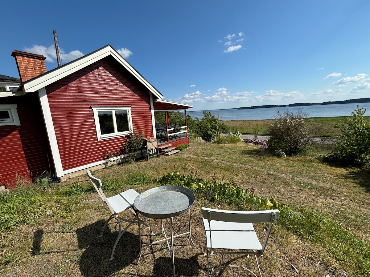 A red wooden house is seen from a grassy area, featuring a small round glass table surrounded by two white chairs. A scenic view of a lake is visible in the background, with a clear blue sky and a few clouds enhancing the tranquil atmosphere.