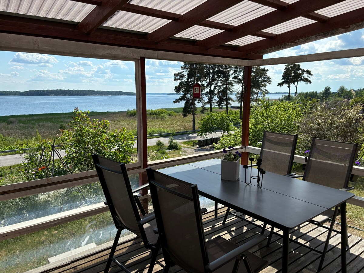 A covered outdoor seating area features a rectangular table surrounded by four chairs. The space offers scenic views of a tranquil body of water, with lush greenery and trees visible in the background under a bright sky.