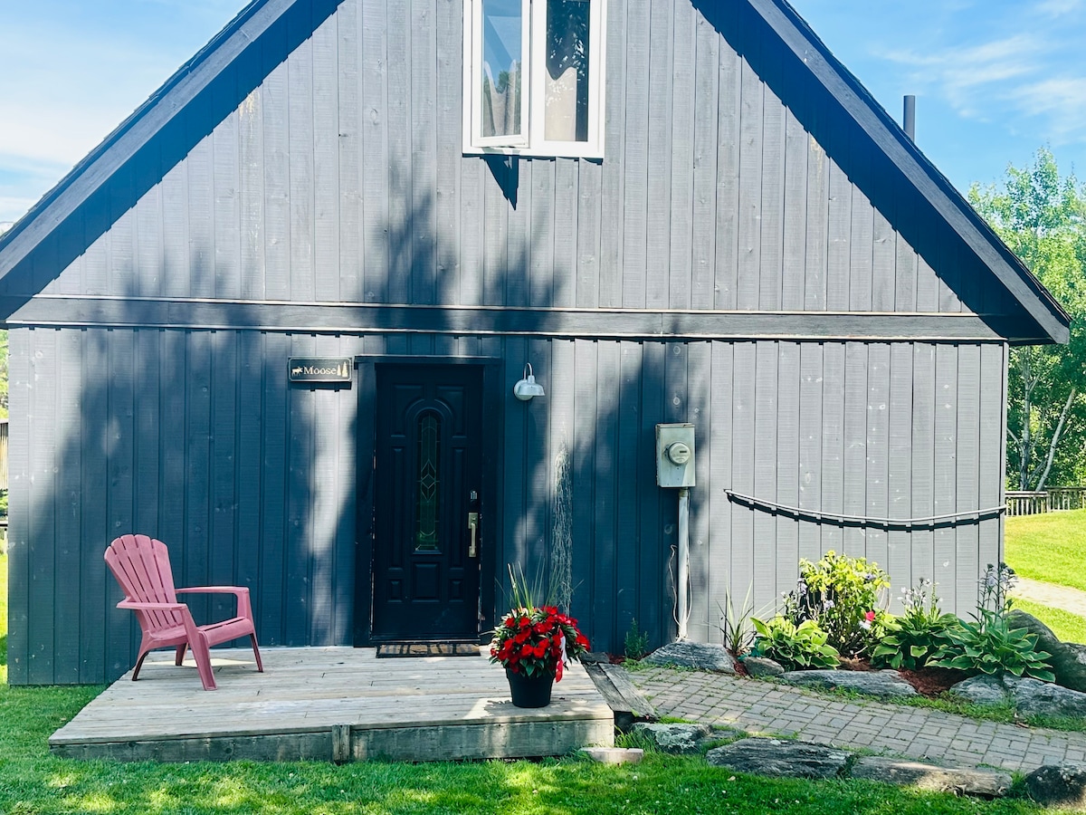 The exterior of the dark-hued A-frame cottage is showcased, featuring a welcoming entrance with a wooden deck and a single pink chair. Colorful flower arrangements enhance the surrounding greenery and decorative stone pathway, leading up to the door beneath a large window.