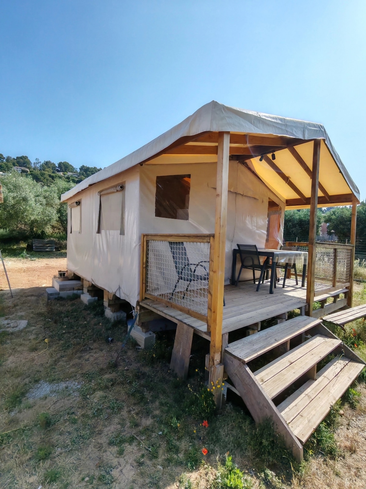A lodge with a light-colored canvas exterior is positioned on a wooden platform. A spacious deck features a small table and two chairs, surrounded by natural greenery. Stairs lead to the entrance, blending the structure with the surrounding landscape of olive trees.