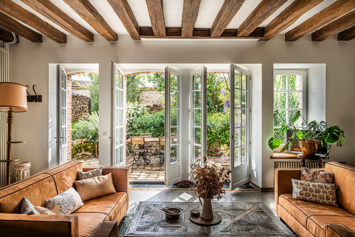 A living area features two large sofas positioned around a coffee table, with natural light streaming through open glass doors. A view of the lush garden is visible, complemented by greenery and a small outdoor table with chairs.