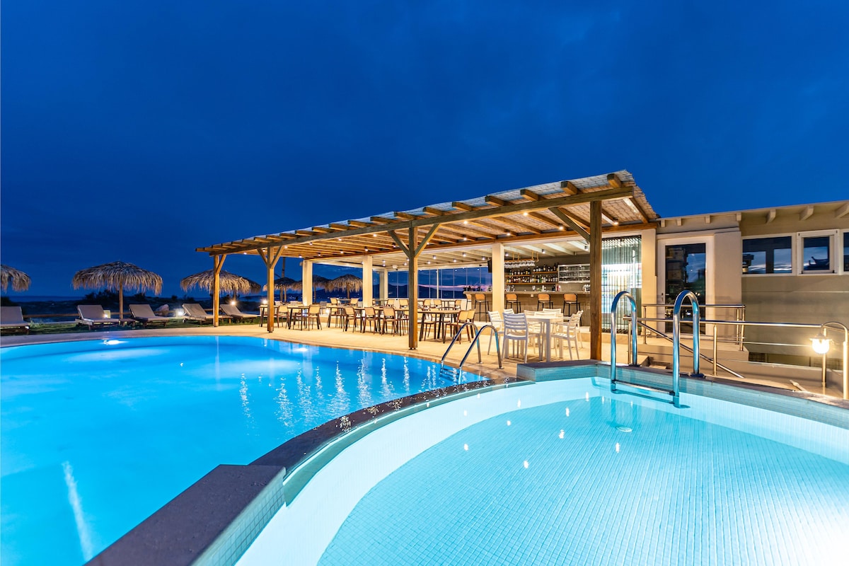 A well-lit swimming pool is surrounded by a wooden pergola and lounge chairs. The shallow area is highlighted, with steps leading into the water. A bar area features seating and overlooks the pool, while umbrellas provide shade nearby.