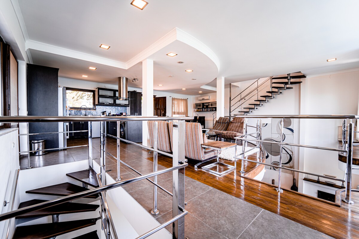 An open-concept living space is revealed with sleek design elements. A spiral staircase leads to an upper level, while comfortable seating arrangements meet a modern kitchen area. Natural light floods through large windows, enhancing the spacious layout and polished flooring.