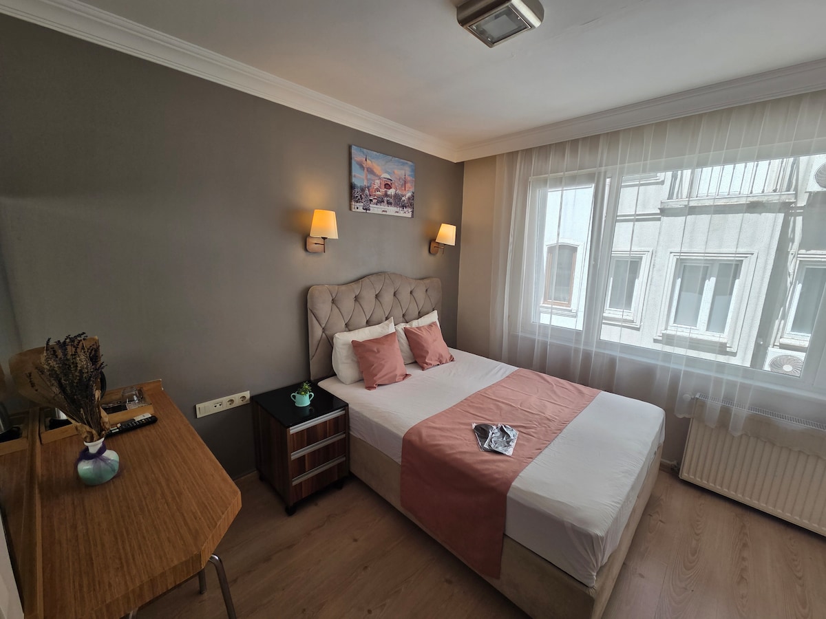 A well-lit bedroom features a spacious bed with a soft headboard, dressed in white linens and an earthy-toned throw. A wooden desk stands beside the bed, complemented by a nightstand. Natural light streams through a large window, creating a calm atmosphere.