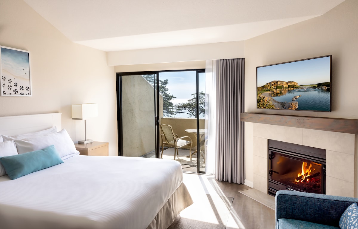 A room features a king-size bed with crisp white linens and a decorative blue pillow. A fireplace is positioned below a wall-mounted television. Floor-to-ceiling glass doors open to a private balcony with views of the ocean, while natural light brightens the space.