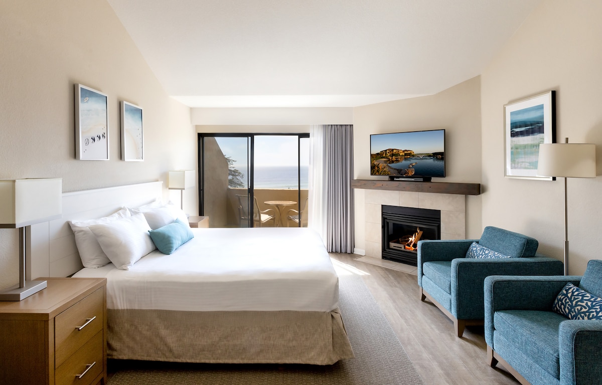 A spacious room features a king size bed with crisp white linens, complemented by two blue chairs. A television is mounted above a fireplace. Large glass doors lead to a private balcony with an ocean view, while natural light illuminates the light-colored walls.