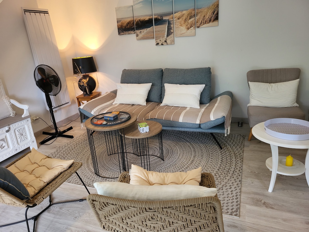 A comfortable living area features a stylish sofa with neutral-colored cushions, accompanied by two woven chairs. A round area rug enhances the space. Multiple tables of varying heights are arranged, and wall art depicting coastal scenes adds a serene touch.