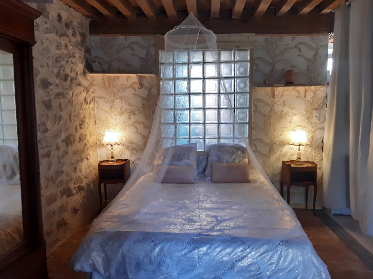A spacious bed is framed by delicate wooden beams and stone walls, illuminated by two bedside lamps. A light canopy drapes over the bed, enhancing the serene atmosphere. Soft, patterned bedding complements the rustic décor, providing a cozy and inviting sleeping area.