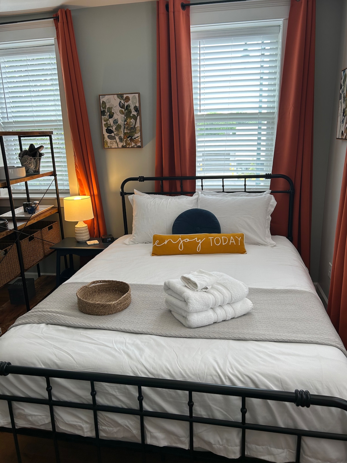 A well-organized sleeping area features a black metal bed with crisp white bedding. An inviting yellow accent pillow rests at the head of the bed. Natural light filters through white blinds, illuminating the orange drapes and a nearby nightstand with a lamp and decorative elements.