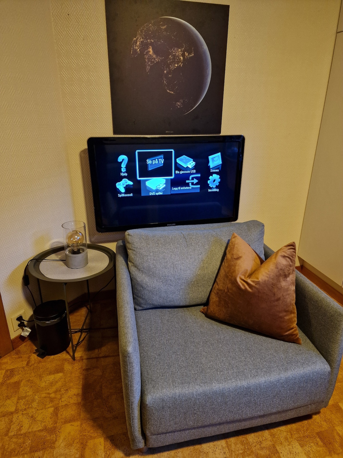 A cozy seating area features a comfortable chair adorned with a brown cushion. Above the chair, a flat-screen TV is mounted, displaying various icons. A small side table with a decorative item and a wastebasket is positioned nearby.