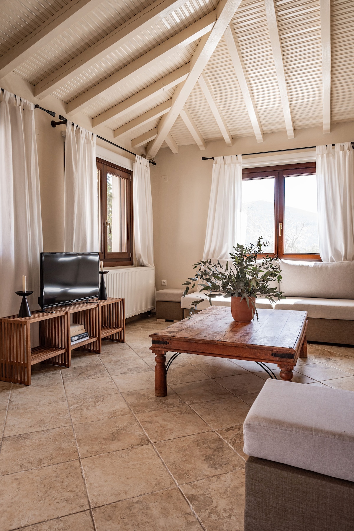 A bright living area features a high, sloped ceiling with wooden beams. Light-colored curtains frame a window, allowing sunlight to illuminate the space. A wooden coffee table with a potted plant sits in the center, complemented by a sectional couch and a television on a simple stand.