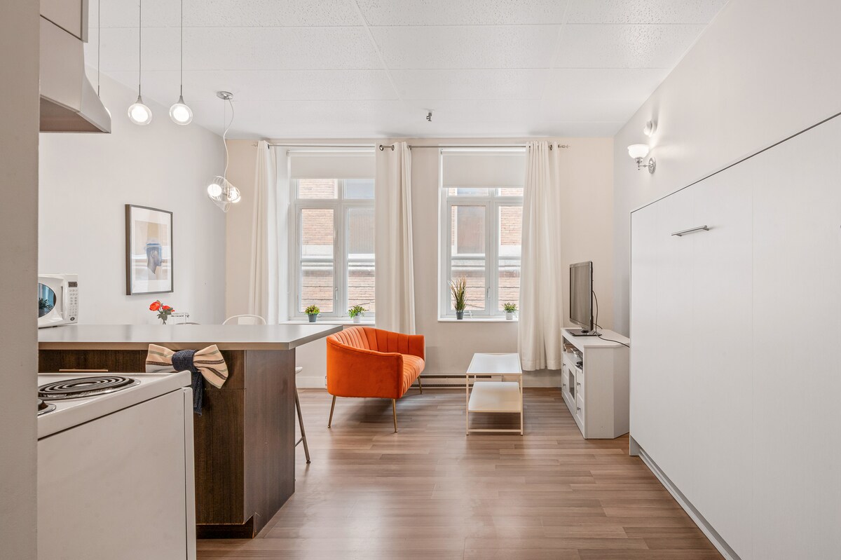 A bright loft space features large windows that allow natural light to illuminate the area. An orange chair adds a pop of color next to a coffee table, while a minimalist entertainment unit holds a TV. The kitchen area is equipped with modern appliances and sleek cabinetry.