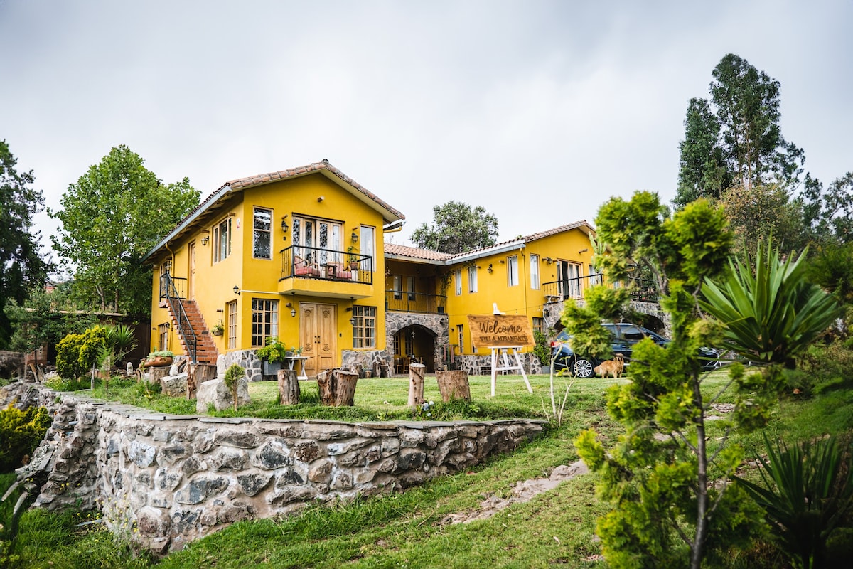 A bright yellow villa is surrounded by lush greenery and stone pathways. The two-story structure features balconies and large windows. Natural stone and wood elements are integrated into the landscape, promoting a rustic charm in a tranquil setting.