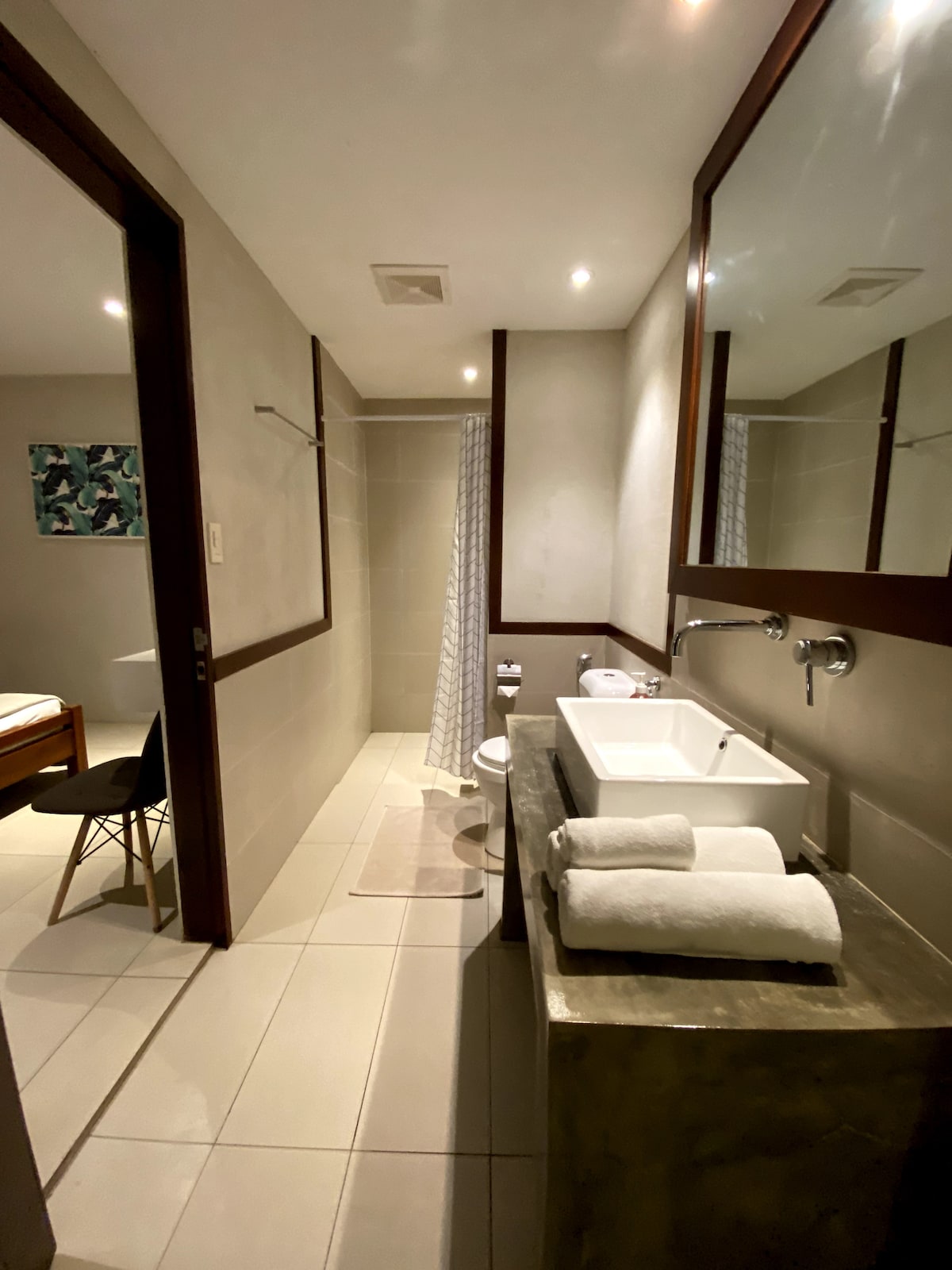 The bathroom is equipped with modern fixtures and has a spacious shower area. A sink with a stone countertop is displayed alongside neatly arranged towels. The design features neutral tones and large mirrors, creating an open and airy feel.