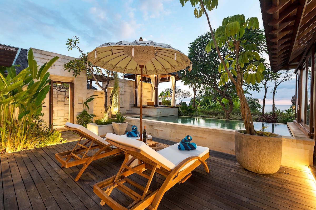 Reclining sunbeds are positioned elegantly on a wooden deck, accompanied by a decorative umbrella. Lush tropical greenery frames the scene, and a private infinity swimming pool is visible in the background, reflecting the serene atmosphere of the outdoor space.