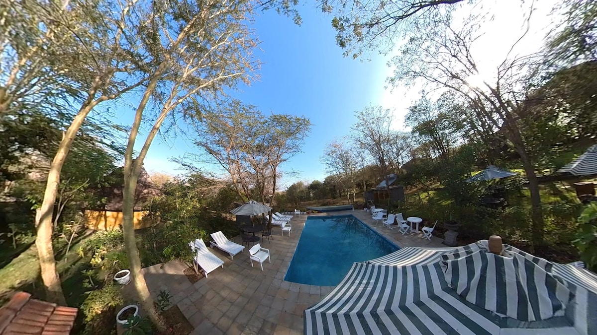 The outdoor pool area features a clear blue swimming pool surrounded by sun loungers and shaded gazebos. Lush greenery and trees provide a natural backdrop, enhancing the tranquil atmosphere. The area is designed for relaxation and enjoyment, inviting guests to unwind in a serene setting.