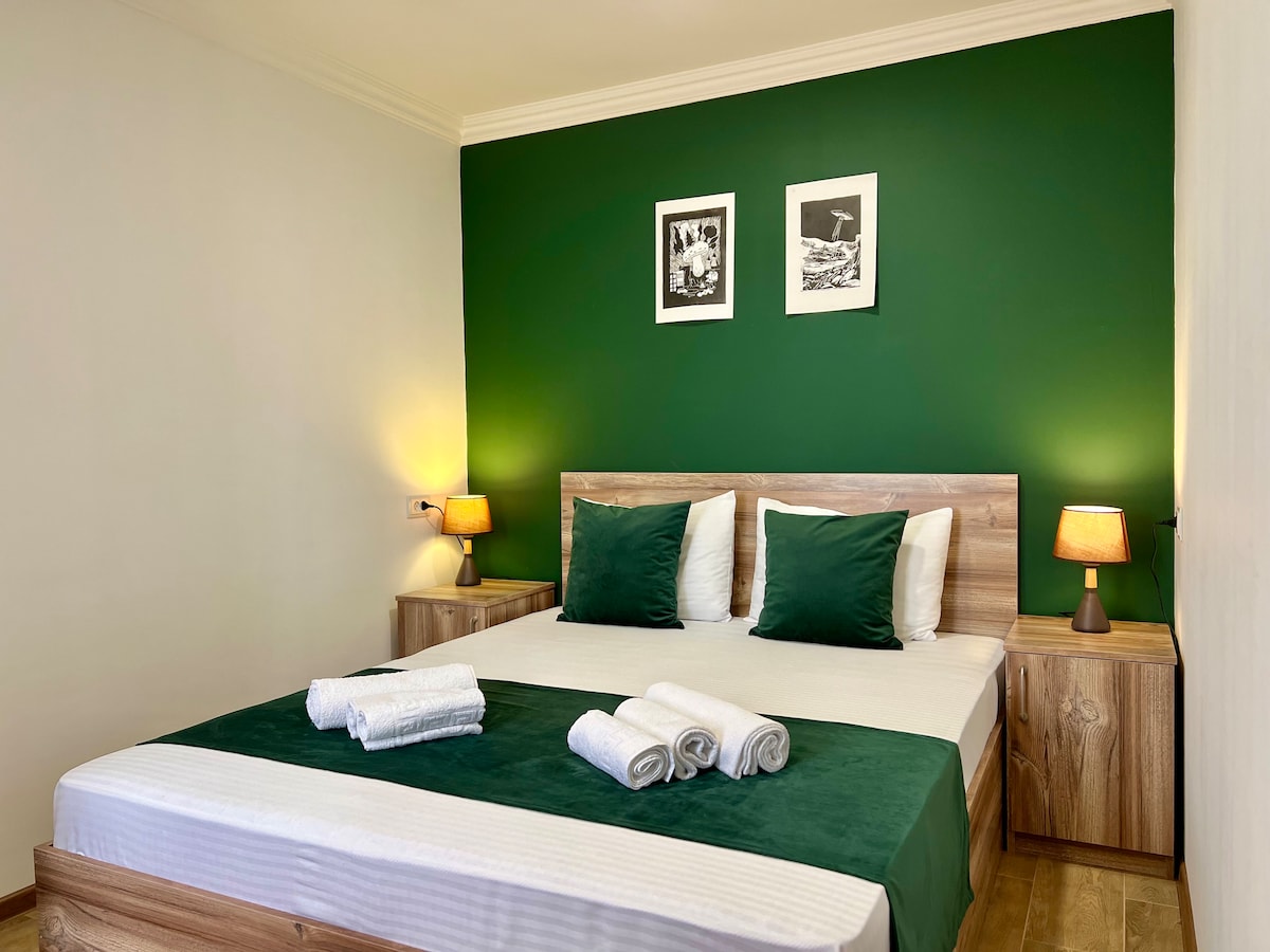 A spacious bedroom is presented with a green accent wall. A large bed is adorned with white linens and a matching green blanket. Nightstands with warm lamps frame the bed. Two framed pictures are hung above the headboard, contributing to a cozy atmosphere.