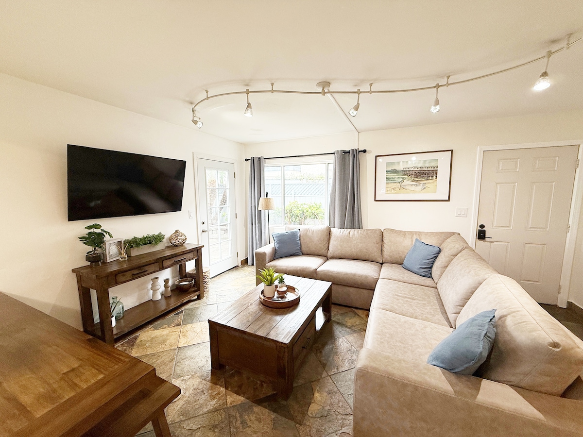 A spacious living area is highlighted by a large sectional sofa, which faces a wall-mounted television. Natural light enters through a sliding glass door, adorned with flowing gray curtains. A wooden coffee table occupies the center, while a decorative plant adds a touch of greenery.