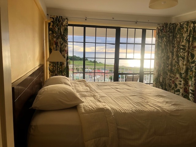 A spacious bedroom features a large bed with a light-colored quilt. Floor-to-ceiling windows offer a scenic view of the outdoors, framed by floral-patterned curtains. Soft lighting from a pendant light adds a gentle brightness to the room.