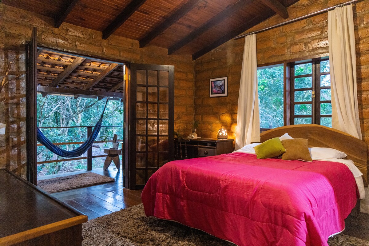 A cozy bedroom features a spacious bed dressed in pink linens, with natural wood accents and warm lighting. Large windows open to a balcony, offering views of the surrounding greenery. A hammock and wooden table are visible outside, inviting relaxation and connection with nature.