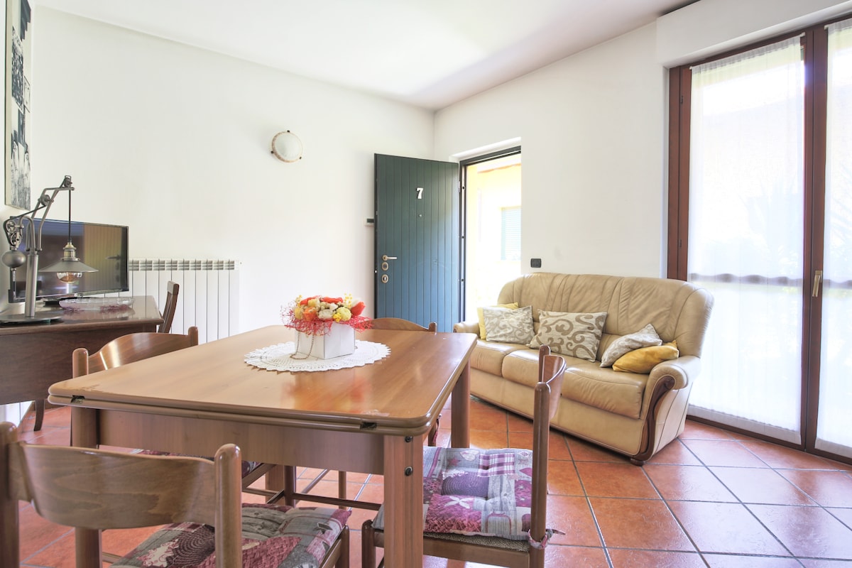 A dining area is displayed with a wooden table accompanied by four chairs. A comfortable sofa with decorative cushions sits nearby. Natural light streams in through large windows, and a door with a number is visible in the background.