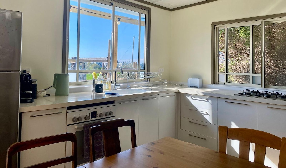 A bright, newly renovated kitchen features a window that allows natural light to fill the space. An inviting wooden dining table is visible, accompanied by several wooden chairs. Modern appliances include a stove, refrigerator, and countertop appliances, enhancing the functional design of the area.