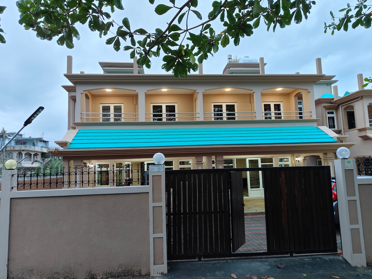 The exterior of the villa is showcased, featuring a two-story structure with a distinctive turquoise roof. A gated entrance provides security, and a paved driveway leads to the main entrance. Lush greenery is seen nearby, contributing to the welcoming environment.