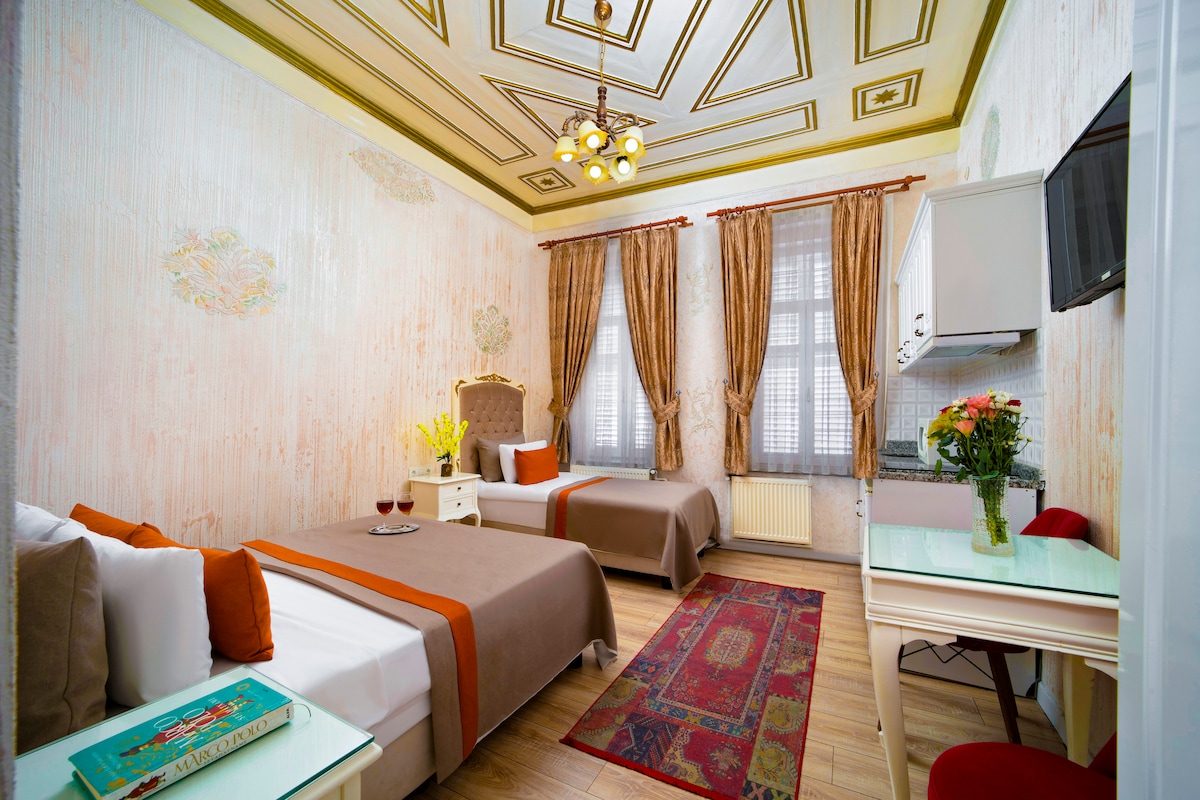 A stylish guest room features two beds with coordinating brown and orange bedding. Natural light enters through large windows adorned with elegant curtains. A small desk is positioned near a wall-mounted television, and a floral arrangement adds a touch of color to the space.