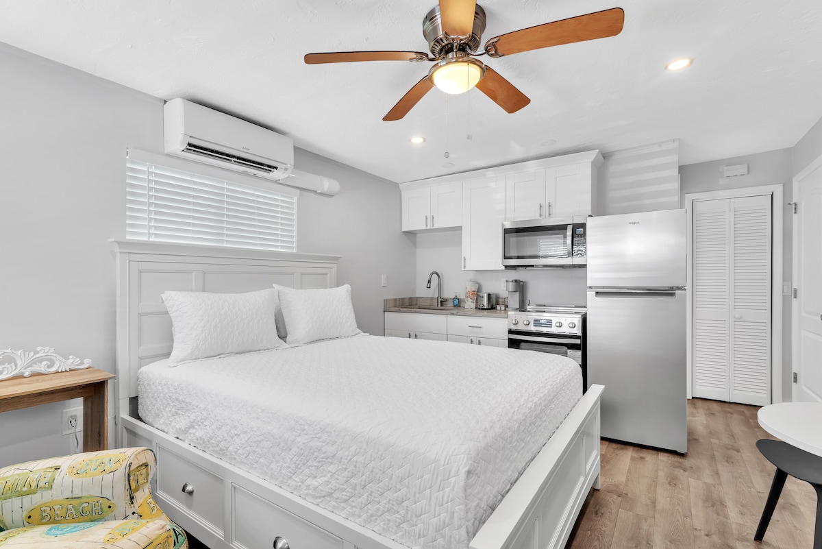 A spacious bedroom is complemented by a king-sized bed with a light quilt. The room features a ceiling fan, air conditioning unit, and a well-equipped kitchen area with a refrigerator, stove, and sink. Natural light enters through the window, enhancing the bright decor.