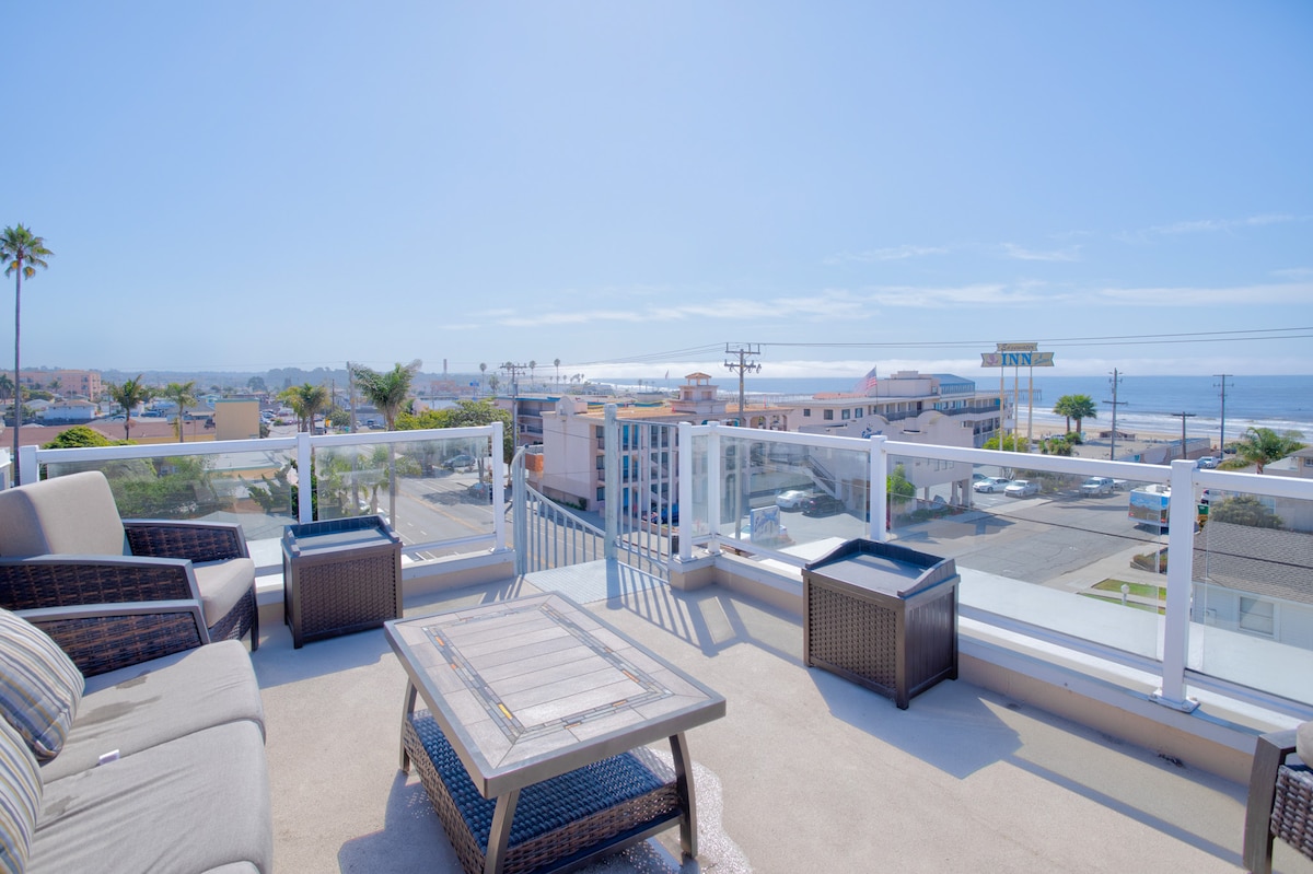 A rooftop lounge area is presented with comfortable seating and a table, offering expansive views of the ocean and surrounding area. The clear blue sky and gentle waves can be seen in the distance, providing a serene backdrop for relaxation.