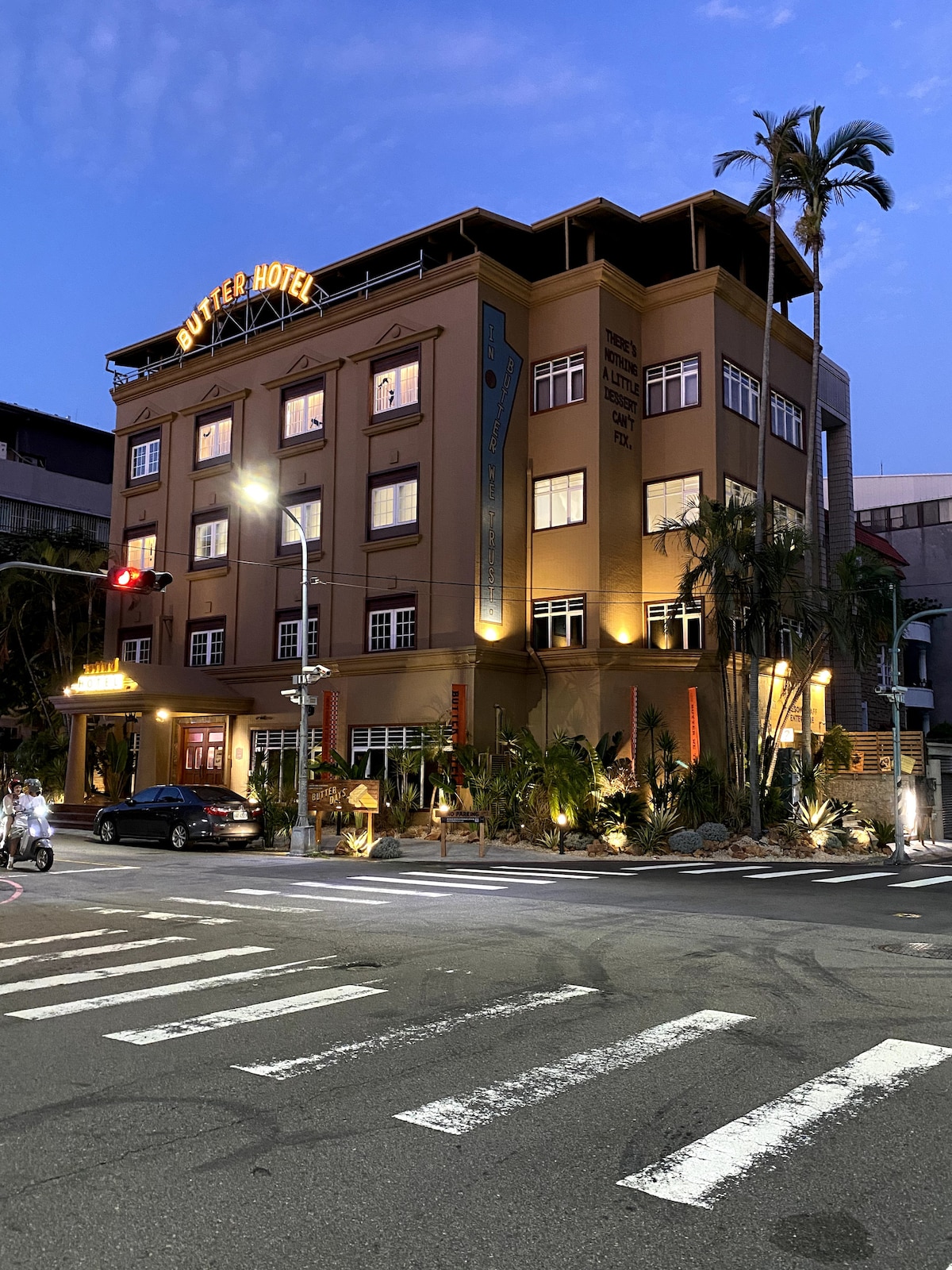 The exterior of Butter Hotel is captured at dusk, showcasing its distinctive architecture illuminated by soft lighting. Lush greenery surrounds the entrance, enhancing the welcoming appearance. Streetlights and traffic signals indicate a lively urban environment, while the building's warm colors contrast with the evening sky.