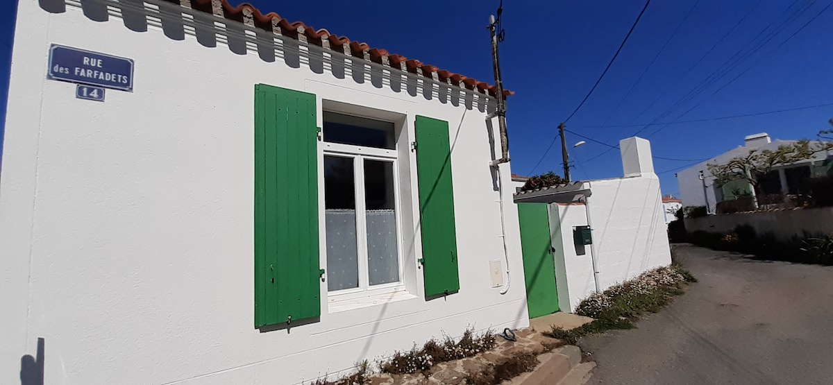 A single-story house is showcased, featuring white walls and green shutters. A gentle slope leads to a small entrance, flanked by a pathway. The roof is adorned with traditional tiles, while the surroundings include sparse vegetation and a clear blue sky above.
