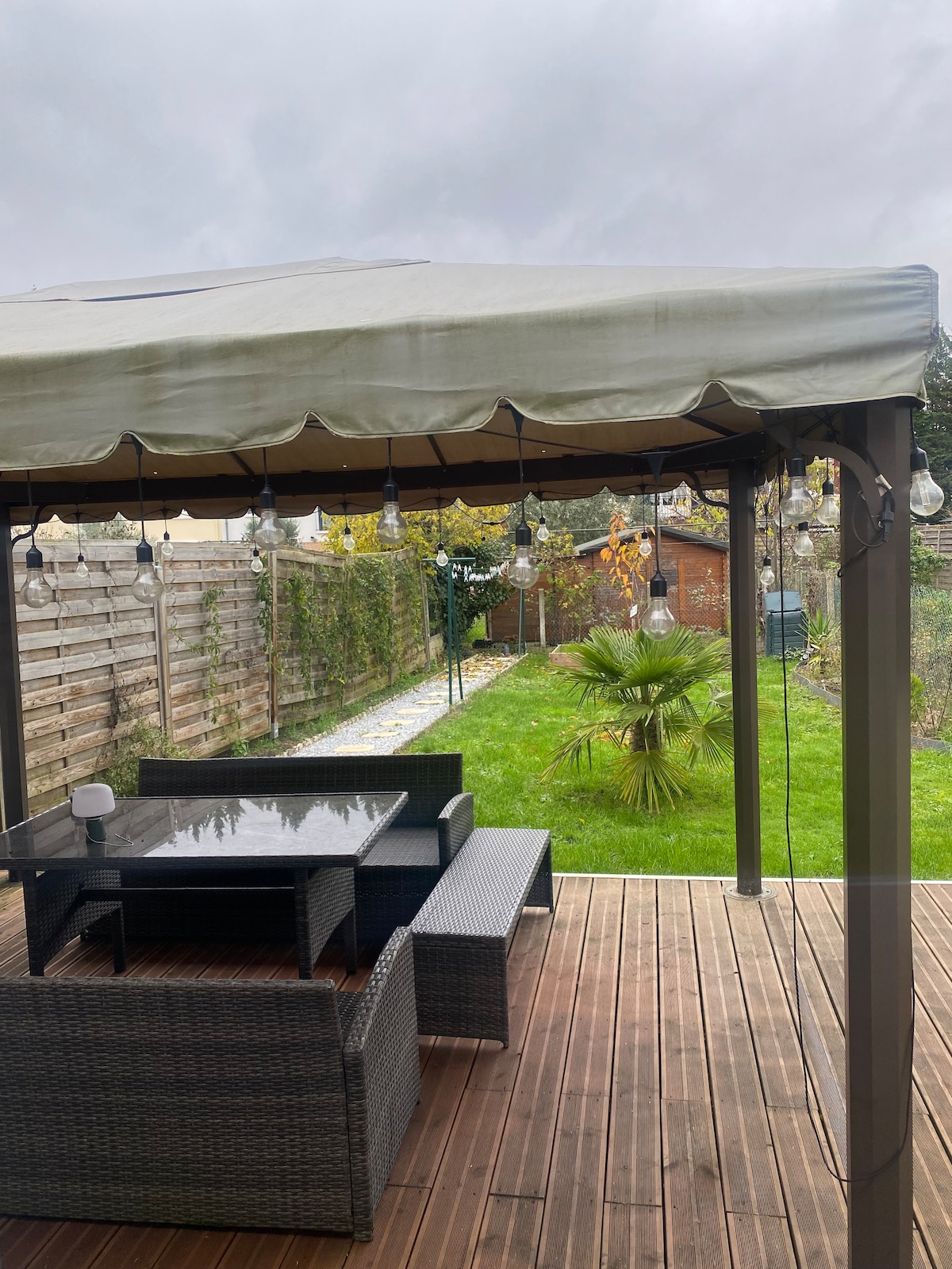 An outdoor seating area is presented under a canopy, with a dark wicker dining set positioned on a wooden deck. The surrounding garden features a lush green lawn and decorative plants, enhancing the tranquil setting. String lights are visible, providing soft illumination.