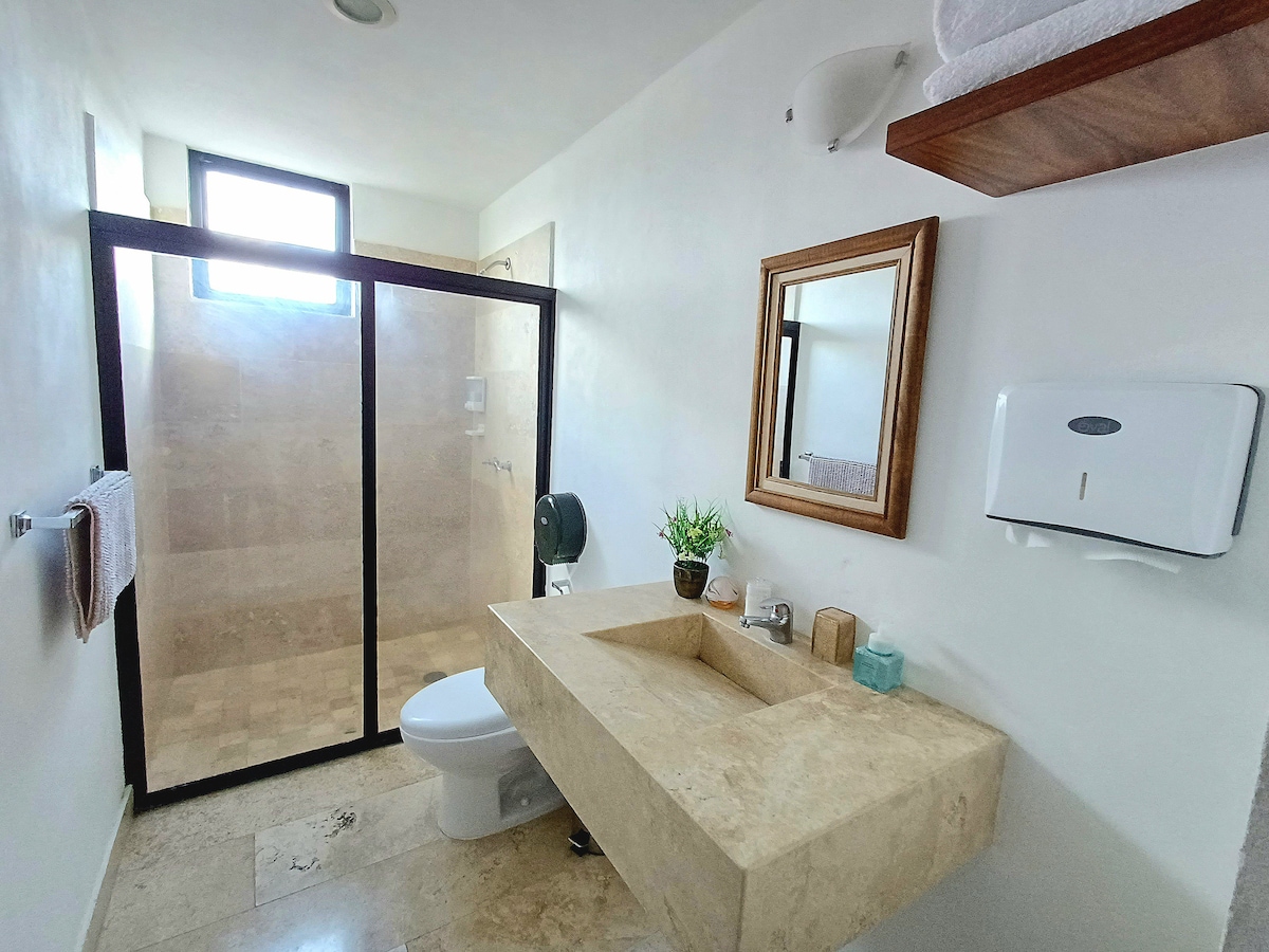 The bathroom features natural stone finishes, with a spacious countertop and a large mirror above the sink. A glass-enclosed shower is visible, along with a toilet and essential toiletries. Natural light enters through a small window, creating a bright and clean environment.