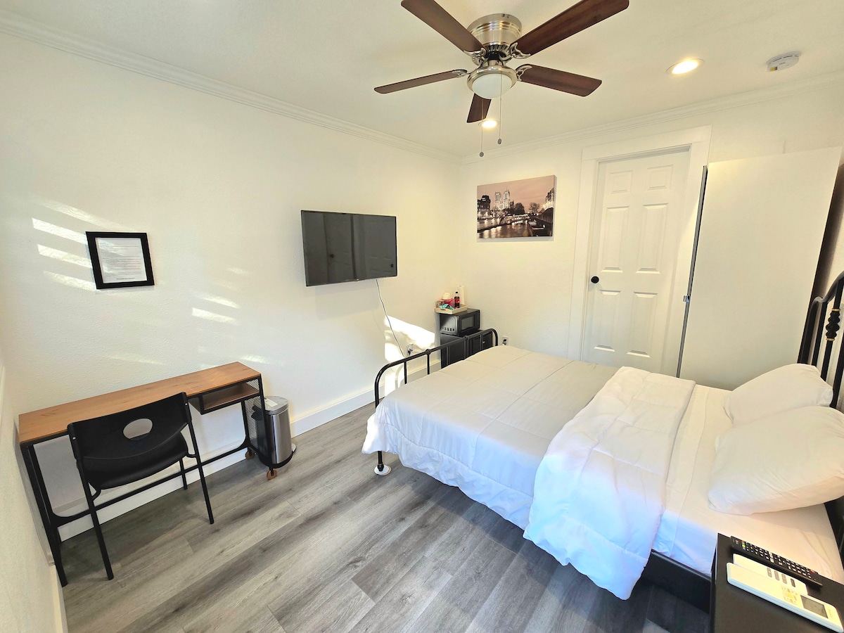 A bright and airy private master suite features a spacious bed with crisp white linens. A television is mounted on the wall across from the bed, and a small desk and chair are placed against one wall. Natural light fills the room through partially opened blinds.