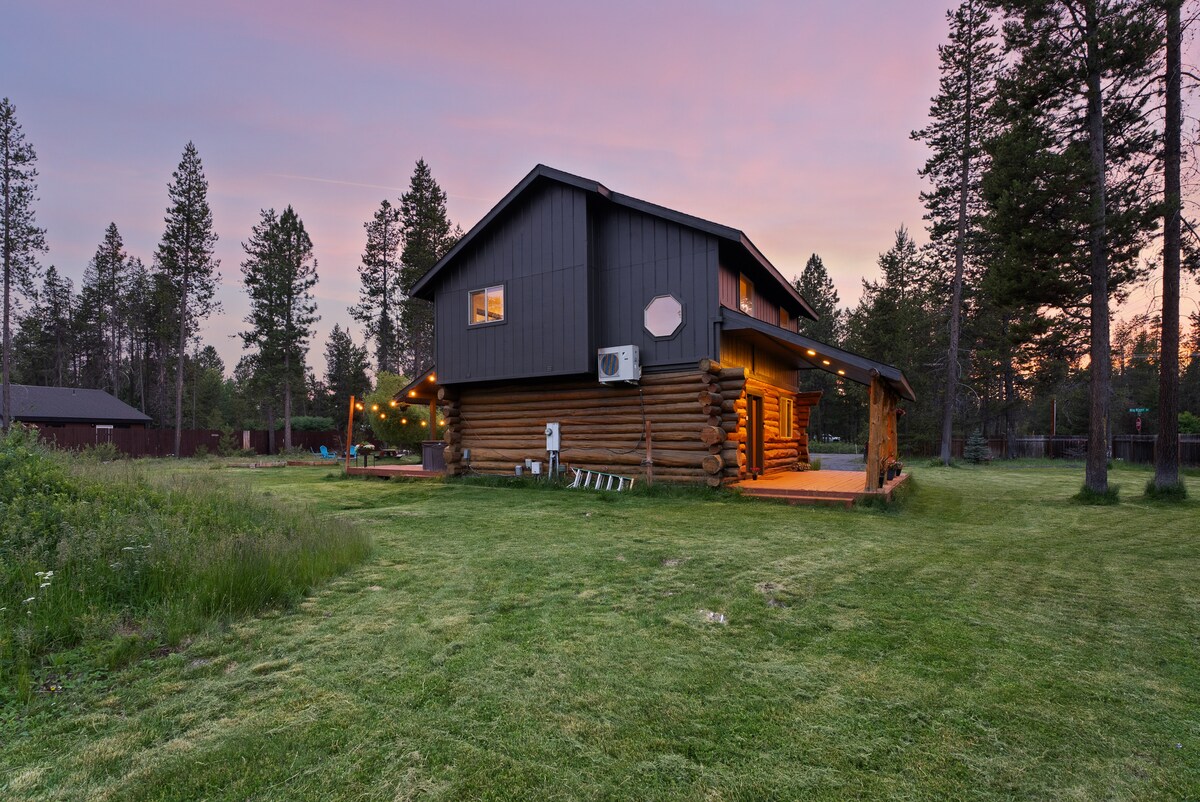 The exterior of a modern cabin showcases a mix of log and contemporary construction under a vibrant twilight sky. A spacious back porch is visible, surrounded by a well-maintained yard with tall grass and scattered pine trees, providing a serene outdoor setting.