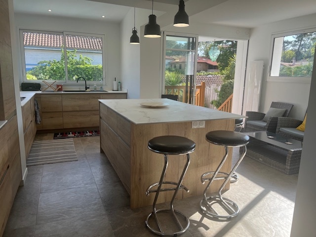 A modern kitchen is displayed with a central island featuring seating on two chrome stools. Natural light is provided by a large window, revealing a view of greenery outside. Sleek wooden cabinets and countertops enhance the inviting space.